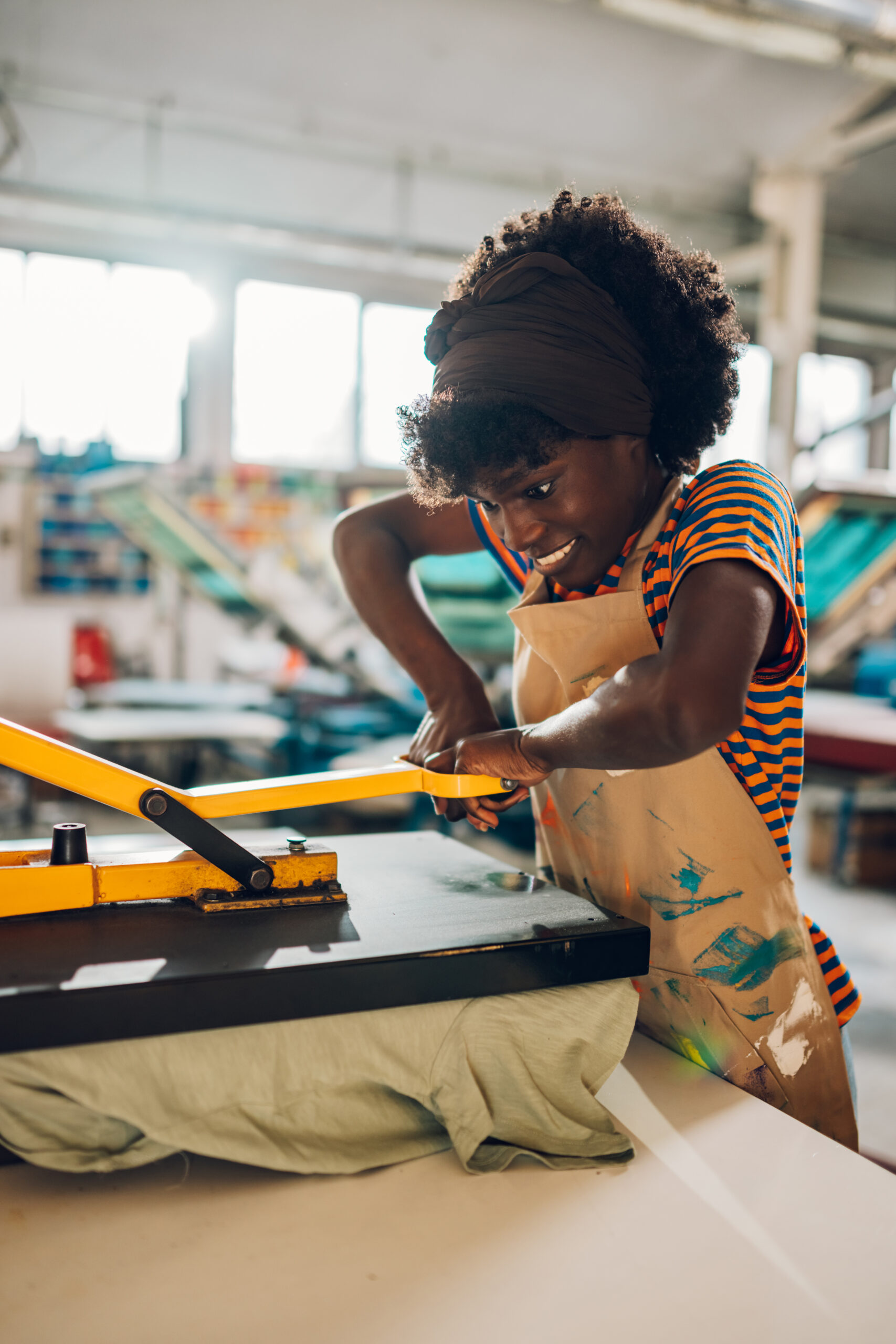 Multicultural printing workshop female worker using heating press on silk screen printed t-shirt at factory. Portrait of african american graphic technologist pressing textile product with heat press